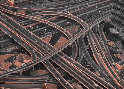 A framed aerial photograph of a sprawling, multi-level highway interchange presented in a selective color style. Dark grey roads weave over and under each other, while the surrounding land is a contrasting copper-brown, creating a dramatic and complex urban pattern. Poster