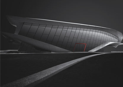 A dramatic black and white photograph of a futuristic, curved building with a dark, reflective facade. A single bright red door provides a striking pop of color. In the foreground, a light gray path curves across a dark landscape, leading the eye towards the structure. The entire image is presented within a silver-colored frame. Decor