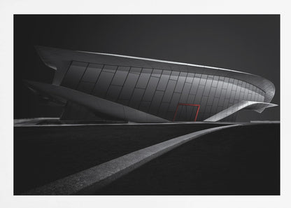 A dramatic black and white photograph of a futuristic, curved building with a dark, reflective facade. A single bright red door provides a striking pop of color. In the foreground, a light gray path curves across a dark landscape, leading the eye towards the structure. The entire image is presented within a silver-colored frame. Decor