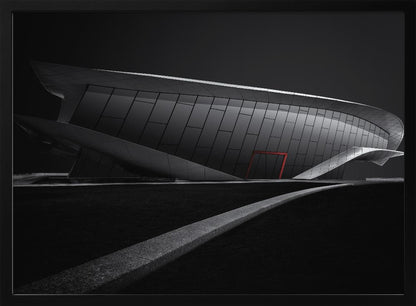 A dramatic black and white photograph of a futuristic, curved building with a dark, reflective facade. A single bright red door provides a striking pop of color. In the foreground, a light gray path curves across a dark landscape, leading the eye towards the structure. The entire image is presented within a silver-colored frame. Decor