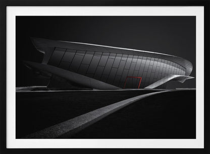 A dramatic black and white photograph of a futuristic, curved building with a dark, reflective facade. A single bright red door provides a striking pop of color. In the foreground, a light gray path curves across a dark landscape, leading the eye towards the structure. The entire image is presented within a silver-colored frame. Decor