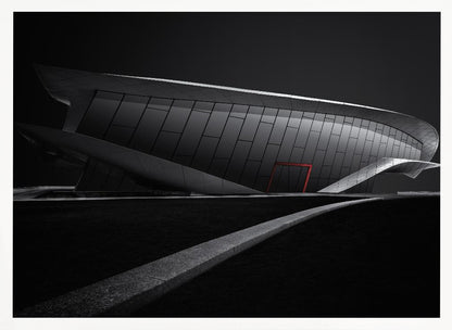 A dramatic black and white photograph of a futuristic, curved building with a dark, reflective facade. A single bright red door provides a striking pop of color. In the foreground, a light gray path curves across a dark landscape, leading the eye towards the structure. The entire image is presented within a silver-colored frame. Decor