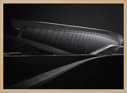 A dramatic black and white photograph of a futuristic, curved building with a dark, reflective facade. A single bright red door provides a striking pop of color. In the foreground, a light gray path curves across a dark landscape, leading the eye towards the structure. The entire image is presented within a silver-colored frame. Decor