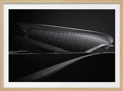 A dramatic black and white photograph of a futuristic, curved building with a dark, reflective facade. A single bright red door provides a striking pop of color. In the foreground, a light gray path curves across a dark landscape, leading the eye towards the structure. The entire image is presented within a silver-colored frame. Decor