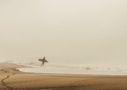 A minimalist photograph of a silhouetted surfer carrying a surfboard, running into the ocean on a foggy morning. The scene has a warm, sepia tone, with footprints visible in the wet sand in the foreground and a vast, misty sky. Wall Art