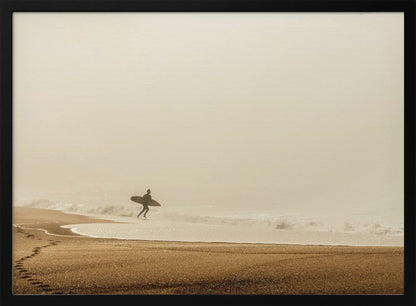 A minimalist photograph of a silhouetted surfer carrying a surfboard, running into the ocean on a foggy morning. The scene has a warm, sepia tone, with footprints visible in the wet sand in the foreground and a vast, misty sky. Wall Art