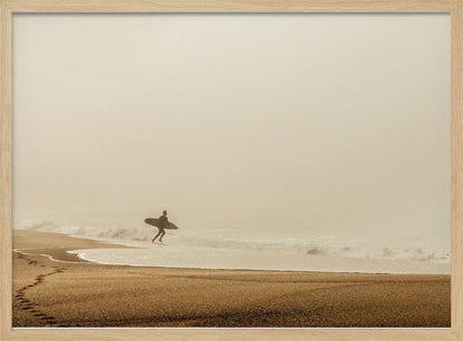 A minimalist photograph of a silhouetted surfer carrying a surfboard, running into the ocean on a foggy morning. The scene has a warm, sepia tone, with footprints visible in the wet sand in the foreground and a vast, misty sky. Wall Art