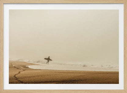 A minimalist photograph of a silhouetted surfer carrying a surfboard, running into the ocean on a foggy morning. The scene has a warm, sepia tone, with footprints visible in the wet sand in the foreground and a vast, misty sky. Wall Art