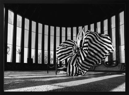 A dramatic black and white photograph of a woman in a voluminous, horizontally striped dress, captured mid-motion with the fabric swirling around her. She stands in a sunlit, circular room with tall, slatted windows that cast long shadows across the floor. Her arms are raised gracefully above her head, creating a striking and artistic pose. Artwork
