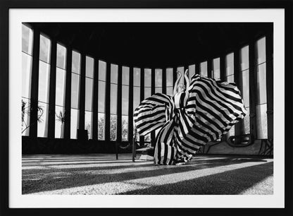 A dramatic black and white photograph of a woman in a voluminous, horizontally striped dress, captured mid-motion with the fabric swirling around her. She stands in a sunlit, circular room with tall, slatted windows that cast long shadows across the floor. Her arms are raised gracefully above her head, creating a striking and artistic pose. Artwork