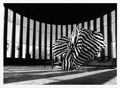 A dramatic black and white photograph of a woman in a voluminous, horizontally striped dress, captured mid-motion with the fabric swirling around her. She stands in a sunlit, circular room with tall, slatted windows that cast long shadows across the floor. Her arms are raised gracefully above her head, creating a striking and artistic pose. Artwork