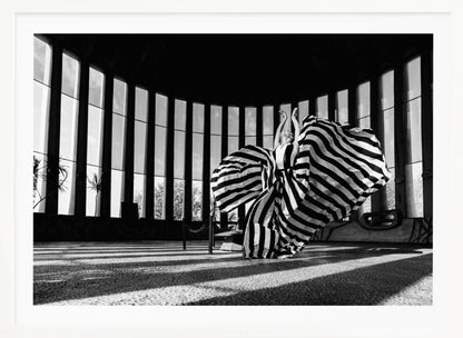 A dramatic black and white photograph of a woman in a voluminous, horizontally striped dress, captured mid-motion with the fabric swirling around her. She stands in a sunlit, circular room with tall, slatted windows that cast long shadows across the floor. Her arms are raised gracefully above her head, creating a striking and artistic pose. Artwork