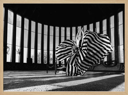 A dramatic black and white photograph of a woman in a voluminous, horizontally striped dress, captured mid-motion with the fabric swirling around her. She stands in a sunlit, circular room with tall, slatted windows that cast long shadows across the floor. Her arms are raised gracefully above her head, creating a striking and artistic pose. Artwork