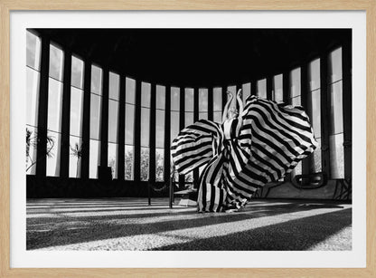 A dramatic black and white photograph of a woman in a voluminous, horizontally striped dress, captured mid-motion with the fabric swirling around her. She stands in a sunlit, circular room with tall, slatted windows that cast long shadows across the floor. Her arms are raised gracefully above her head, creating a striking and artistic pose. Artwork