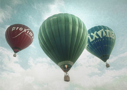 A low-angle photograph of three colorful hot air balloons floating in a cloudy blue sky, presented in a silver frame. A large green balloon is in the center, with a red balloon on the left and a dark blue balloon on the right, all featuring logos. Wall Art