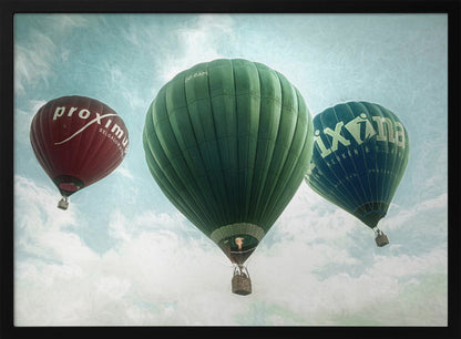 A low-angle photograph of three colorful hot air balloons floating in a cloudy blue sky, presented in a silver frame. A large green balloon is in the center, with a red balloon on the left and a dark blue balloon on the right, all featuring logos. Wall Art