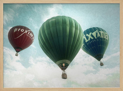A low-angle photograph of three colorful hot air balloons floating in a cloudy blue sky, presented in a silver frame. A large green balloon is in the center, with a red balloon on the left and a dark blue balloon on the right, all featuring logos. Wall Art