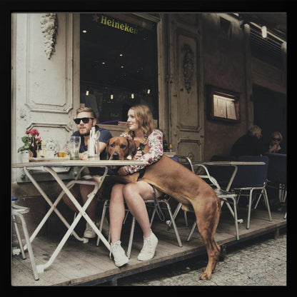 A framed photograph of a young couple sitting at an outdoor cafe. A woman in a floral shirt holds a large brown Rhodesian Ridgeback on her lap, while a man in sunglasses sits beside her. The dog stands with its front paws on the woman's lap and back paws on the wooden deck. The scene has a muted, warm tone. Poster