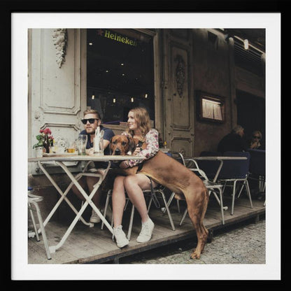 A framed photograph of a young couple sitting at an outdoor cafe. A woman in a floral shirt holds a large brown Rhodesian Ridgeback on her lap, while a man in sunglasses sits beside her. The dog stands with its front paws on the woman's lap and back paws on the wooden deck. The scene has a muted, warm tone. Poster
