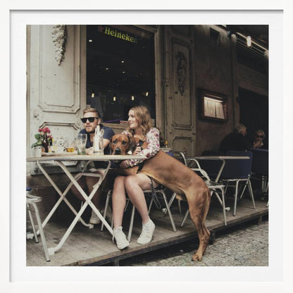 A framed photograph of a young couple sitting at an outdoor cafe. A woman in a floral shirt holds a large brown Rhodesian Ridgeback on her lap, while a man in sunglasses sits beside her. The dog stands with its front paws on the woman's lap and back paws on the wooden deck. The scene has a muted, warm tone. Poster