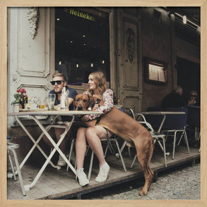 A framed photograph of a young couple sitting at an outdoor cafe. A woman in a floral shirt holds a large brown Rhodesian Ridgeback on her lap, while a man in sunglasses sits beside her. The dog stands with its front paws on the woman's lap and back paws on the wooden deck. The scene has a muted, warm tone. Poster