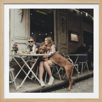 A framed photograph of a young couple sitting at an outdoor cafe. A woman in a floral shirt holds a large brown Rhodesian Ridgeback on her lap, while a man in sunglasses sits beside her. The dog stands with its front paws on the woman's lap and back paws on the wooden deck. The scene has a muted, warm tone. Poster