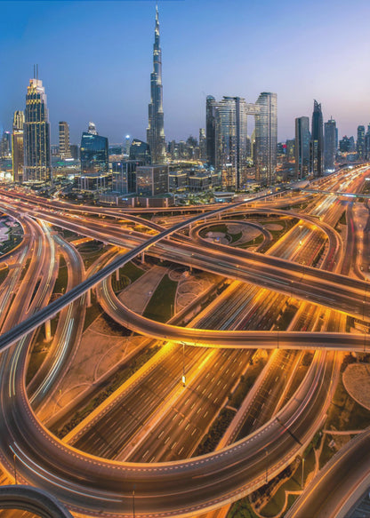 A long-exposure, aerial photograph of the Dubai skyline at dusk. The iconic Burj Khalifa stands tall in the center, surrounded by other illuminated skyscrapers against a deep blue twilight sky. In the foreground, a complex web of highways and overpasses glows with the orange and white light trails of moving traffic. Decor