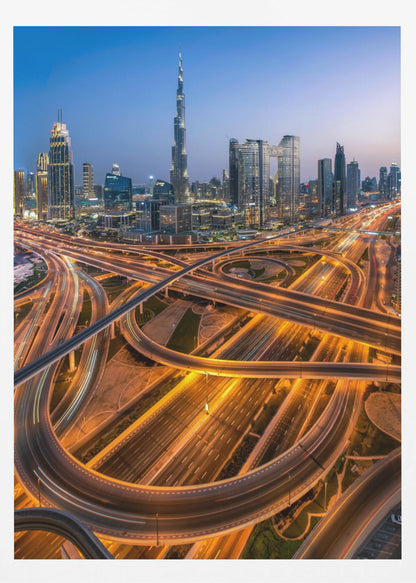 A long-exposure, aerial photograph of the Dubai skyline at dusk. The iconic Burj Khalifa stands tall in the center, surrounded by other illuminated skyscrapers against a deep blue twilight sky. In the foreground, a complex web of highways and overpasses glows with the orange and white light trails of moving traffic. Decor