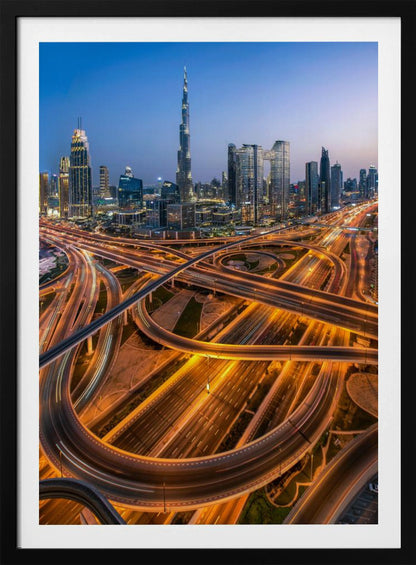A long-exposure, aerial photograph of the Dubai skyline at dusk. The iconic Burj Khalifa stands tall in the center, surrounded by other illuminated skyscrapers against a deep blue twilight sky. In the foreground, a complex web of highways and overpasses glows with the orange and white light trails of moving traffic. Decor