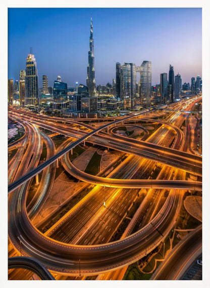A long-exposure, aerial photograph of the Dubai skyline at dusk. The iconic Burj Khalifa stands tall in the center, surrounded by other illuminated skyscrapers against a deep blue twilight sky. In the foreground, a complex web of highways and overpasses glows with the orange and white light trails of moving traffic. Decor