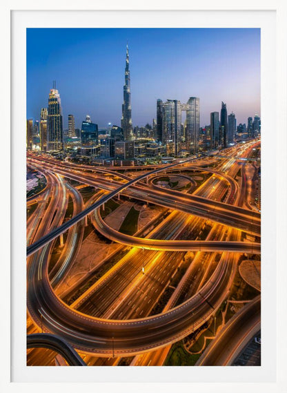 A long-exposure, aerial photograph of the Dubai skyline at dusk. The iconic Burj Khalifa stands tall in the center, surrounded by other illuminated skyscrapers against a deep blue twilight sky. In the foreground, a complex web of highways and overpasses glows with the orange and white light trails of moving traffic. Decor