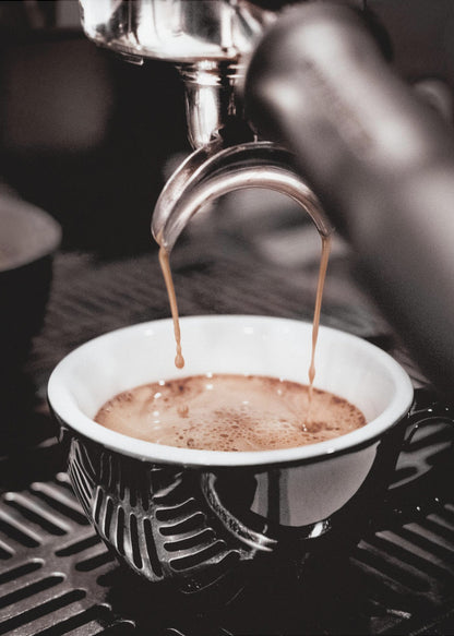 A close-up, desaturated photograph of an espresso machine pouring two streams of rich brown coffee from a chrome portafilter into an ornate black and white cup. Poster