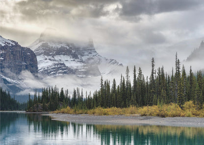 A tranquil mountain landscape featuring a still, teal lake reflecting a dense evergreen forest along its shore. In the background, majestic snow-covered peaks are shrouded in mist and dramatic gray clouds. Wall Art