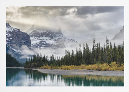 A tranquil mountain landscape featuring a still, teal lake reflecting a dense evergreen forest along its shore. In the background, majestic snow-covered peaks are shrouded in mist and dramatic gray clouds. Wall Art