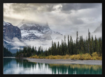 A tranquil mountain landscape featuring a still, teal lake reflecting a dense evergreen forest along its shore. In the background, majestic snow-covered peaks are shrouded in mist and dramatic gray clouds. Wall Art
