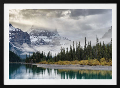 A tranquil mountain landscape featuring a still, teal lake reflecting a dense evergreen forest along its shore. In the background, majestic snow-covered peaks are shrouded in mist and dramatic gray clouds. Wall Art