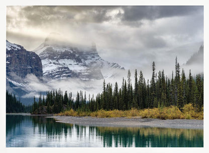A tranquil mountain landscape featuring a still, teal lake reflecting a dense evergreen forest along its shore. In the background, majestic snow-covered peaks are shrouded in mist and dramatic gray clouds. Wall Art