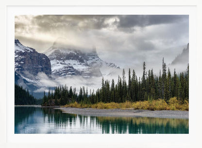 A tranquil mountain landscape featuring a still, teal lake reflecting a dense evergreen forest along its shore. In the background, majestic snow-covered peaks are shrouded in mist and dramatic gray clouds. Wall Art
