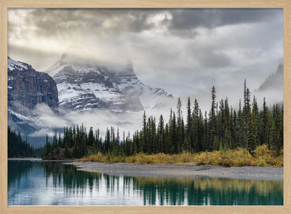 A tranquil mountain landscape featuring a still, teal lake reflecting a dense evergreen forest along its shore. In the background, majestic snow-covered peaks are shrouded in mist and dramatic gray clouds. Wall Art