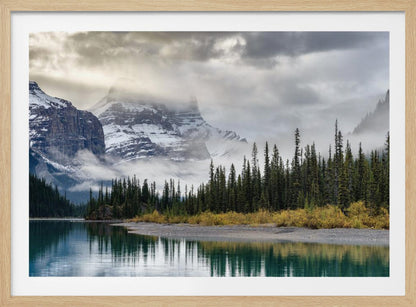 A tranquil mountain landscape featuring a still, teal lake reflecting a dense evergreen forest along its shore. In the background, majestic snow-covered peaks are shrouded in mist and dramatic gray clouds. Wall Art