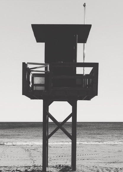 A high-contrast, black and white photograph of a silhouetted lifeguard tower standing on a sandy beach. The tower is centrally framed against a bright, empty sky, with the ocean and horizon visible in the background. Wall Art