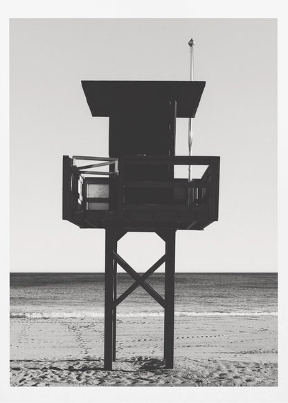 A high-contrast, black and white photograph of a silhouetted lifeguard tower standing on a sandy beach. The tower is centrally framed against a bright, empty sky, with the ocean and horizon visible in the background. Wall Art