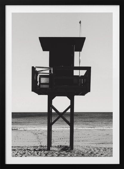 A high-contrast, black and white photograph of a silhouetted lifeguard tower standing on a sandy beach. The tower is centrally framed against a bright, empty sky, with the ocean and horizon visible in the background. Wall Art