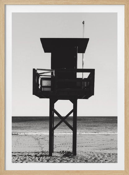 A high-contrast, black and white photograph of a silhouetted lifeguard tower standing on a sandy beach. The tower is centrally framed against a bright, empty sky, with the ocean and horizon visible in the background. Wall Art