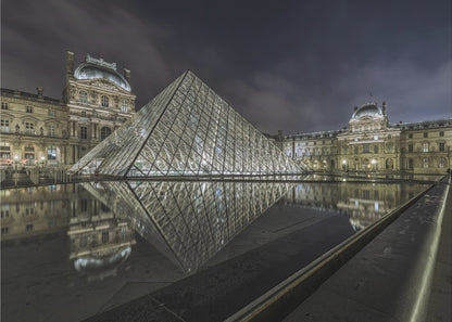 A dramatic night photograph of the illuminated Louvre Pyramid in Paris, its glass structure glowing against a dark, cloudy sky. The historic Louvre Palace is visible in the background, and the entire scene is perfectly reflected in the still water of a foreground pool. The image is presented within a silver frame. Artwork