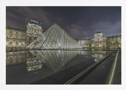 A dramatic night photograph of the illuminated Louvre Pyramid in Paris, its glass structure glowing against a dark, cloudy sky. The historic Louvre Palace is visible in the background, and the entire scene is perfectly reflected in the still water of a foreground pool. The image is presented within a silver frame. Artwork