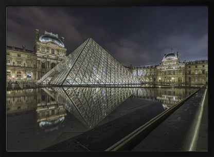 A dramatic night photograph of the illuminated Louvre Pyramid in Paris, its glass structure glowing against a dark, cloudy sky. The historic Louvre Palace is visible in the background, and the entire scene is perfectly reflected in the still water of a foreground pool. The image is presented within a silver frame. Artwork