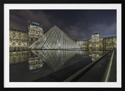 A dramatic night photograph of the illuminated Louvre Pyramid in Paris, its glass structure glowing against a dark, cloudy sky. The historic Louvre Palace is visible in the background, and the entire scene is perfectly reflected in the still water of a foreground pool. The image is presented within a silver frame. Artwork