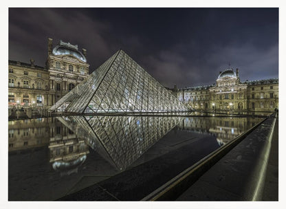 A dramatic night photograph of the illuminated Louvre Pyramid in Paris, its glass structure glowing against a dark, cloudy sky. The historic Louvre Palace is visible in the background, and the entire scene is perfectly reflected in the still water of a foreground pool. The image is presented within a silver frame. Artwork