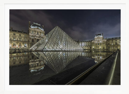 A dramatic night photograph of the illuminated Louvre Pyramid in Paris, its glass structure glowing against a dark, cloudy sky. The historic Louvre Palace is visible in the background, and the entire scene is perfectly reflected in the still water of a foreground pool. The image is presented within a silver frame. Artwork
